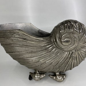 Op de afbeelding: Een zilveren metalen schelpvormige plantenbak met een getextureerd oppervlak en drie poten. De plantenbak is gevormd als een schelp en heeft een spiraalvormig ontwerp.
