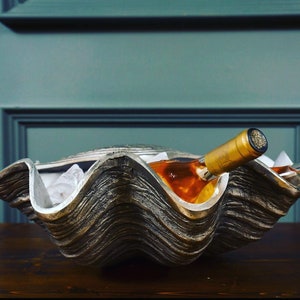 May include: A large, decorative clam shell wine chiller. The shell is a mix of silver and bronze, with a textured surface. Two wine bottles rest inside, surrounded by ice.