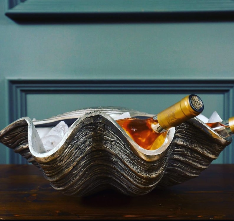 May include: A large, decorative clam shell wine chiller. The shell is a mix of silver and bronze, with a textured surface. Two wine bottles rest inside, surrounded by ice.