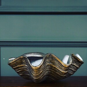 May include: A large, decorative clam shell bowl with a textured, metallic gold and silver finish. The bowl has a wavy, layered design and sits on a dark wooden surface. The background is a muted teal color.