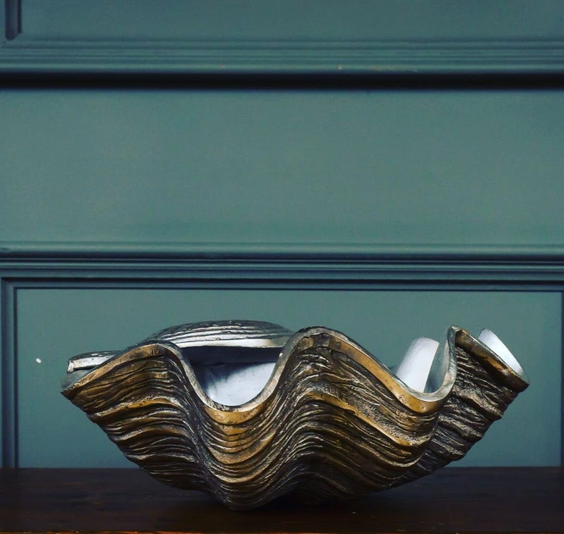 May include: A large, decorative clam shell bowl with a textured, metallic gold and silver finish. The bowl has a wavy, layered design and sits on a dark wooden surface. The background is a muted teal color.