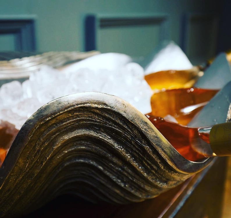 May include: A close-up of a decorative, wave-shaped serving dish with a metallic finish. The dish is filled with ice and a golden-colored liquid. The edges of the dish have a textured, layered appearance.