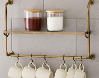 May include: A brass-coloured kitchen shelf with a glass shelf and a row of hooks. Two glass jars with wooden lids sit on the shelf, one filled with a red substance and the other with a white substance. Six white mugs hang from the hooks.