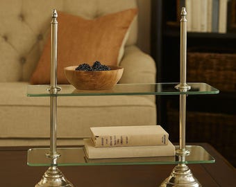 Puede incluir: Una mesa auxiliar de dos niveles con estantes de vidrio y estructura metálica plateada. La mesa tiene dos estantes rectangulares de vidrio. Un cuenco de madera con moras y dos libros están sobre los estantes. La mesa está sobre una superficie marrón oscuro.