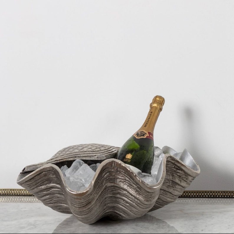 May include: A silver-colored clam shell ice bucket with a bottle of champagne inside. The ice bucket has a textured surface and is filled with ice cubes. The champagne bottle has a gold foil label.