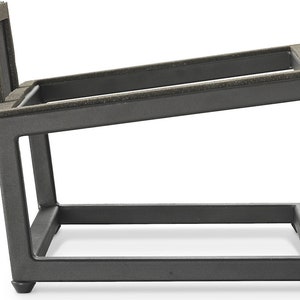 Può includere: Un supporto in metallo grigio scuro con una parte superiore inclinata e una base rettangolare. Il supporto ha una superficie imbottita sulla parte superiore e un design geometrico robusto. Il supporto è progettato per contenere un oggetto.