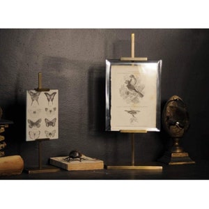 May include: Two brass picture stands displaying a vintage print of birds and a print of butterflies. The stands feature a simple design with a vertical rod and a horizontal base.
