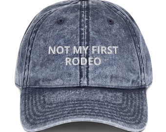 May include: A blue denim baseball cap with the text "NOT MY FIRST RODEO" embroidered in white on the front.