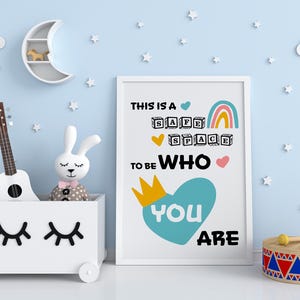 May include: A white framed wall art print featuring the text THIS IS A SAFE SPACE TO BE WHO YOU ARE, decorated with a rainbow, a yellow crown, and a large teal heart. The print is displayed next to a white toy chest with a ukulele and rabbit doll.