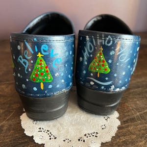 May include: A pair of blue clogs with hand-painted Christmas tree designs. One clog says "Believe" and the other says "Ho Ho Ho". The trees are green with red ornaments and a yellow star. White polka dots and snowflakes are scattered across the clogs.
