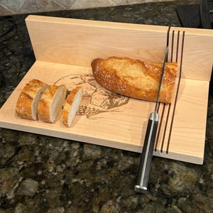 Könnte beinhalten: Ein Holzbrot-Schneidebrett mit integriertem Messerführung und einem dekorativen, gravierten Design. Ein Brotlaib wird auf dem Brett mit einem silbernen Messer geschnitten.