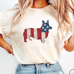 May include: Cream-colored t-shirt featuring a patriotic corgi design. The corgi is depicted with the American flag, showcasing red and white stripes on its body and a blue head with white stars. A casual, comfortable style.
