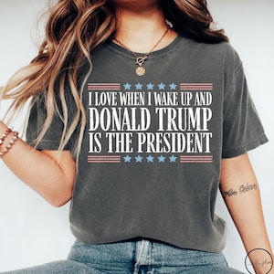 Op de afbeelding: Een donkergrijs T-shirt met een witte en blauwe grafische print met de tekst "Ik hou ervan om wakker te worden en Donald Trump is president". De grafiek bevat ook rode en blauwe strepen en sterren.