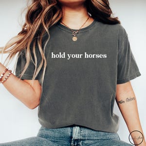 Op de afbeelding: Een donkergrijs T-shirt met de tekst "hold your horses" in wit gedrukt op de voorkant. Het shirt is gemaakt van een zacht, comfortabel materiaal en heeft een ontspannen pasvorm.