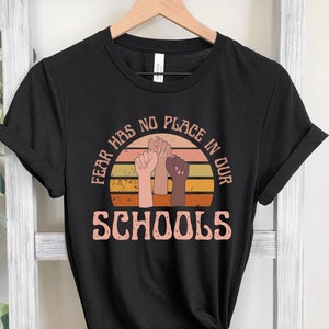 May include: Black t-shirt with a retro design featuring the text "FEAR HAS NO PLACE IN OUR SCHOOLS" in a circular design with raised fists in various skin tones. The design is in shades of orange, yellow, and brown.
