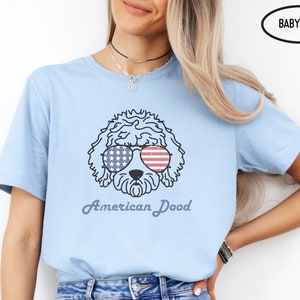 May include: A light blue t-shirt with a cartoon doodle of a dog wearing sunglasses with an American flag design. The text "American Dood" is printed below the dog.