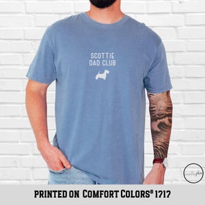 May include: A blue t-shirt with the text "SCOTTIE DAD CLUB" and a white silhouette of a Scottish Terrier. The shirt is printed on Comfort Colors 1212 fabric.