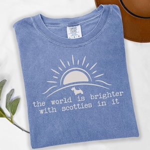 May include: A light blue t-shirt with a white graphic of a sun, a Scottie dog, and the text "the world is brighter with scotties in it." The shirt is a Comfort Colors brand, size L-G-G.
