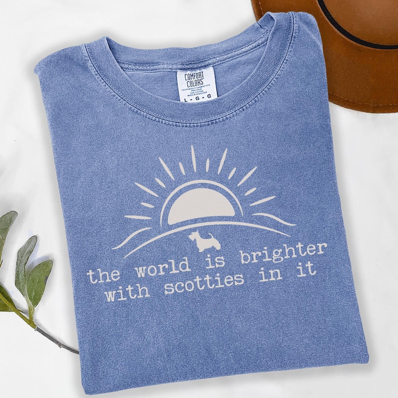 May include: A light blue t-shirt with a white graphic of a sun, a Scottie dog, and the text "the world is brighter with scotties in it." The shirt is a Comfort Colors brand, size L-G-G.