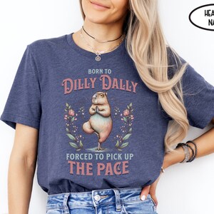 May include: A heather navy blue t-shirt featuring a graphic of a capybara doing yoga. The text on the shirt reads "Born to Dilly Dally Forced to Pick Up The Pace".