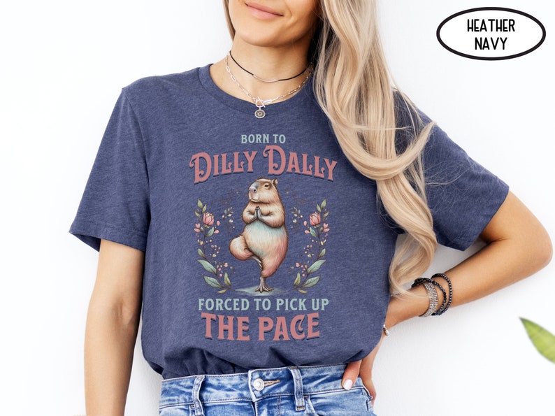 May include: A heather navy blue t-shirt featuring a graphic of a capybara doing yoga. The text on the shirt reads "Born to Dilly Dally Forced to Pick Up The Pace".