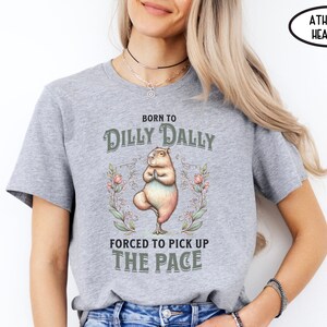 May include: Grey athletic heather t-shirt featuring a graphic of a capybara doing yoga. The text on the shirt reads "Born to Dilly Dally Forced to Pick Up The Pace".