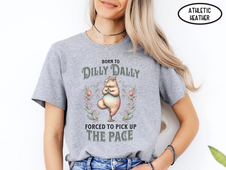 May include: Grey athletic heather t-shirt featuring a graphic of a capybara doing yoga. The text on the shirt reads "Born to Dilly Dally Forced to Pick Up The Pace".