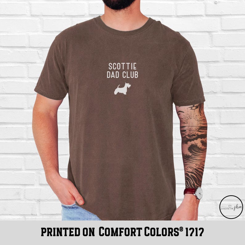May include: A brown t-shirt with the text "SCOTTIE DAD CLUB" and a white Scottie dog silhouette. The phrase "PRINTED ON COMFORT COLORS&reg; 1212" is printed at the bottom of the shirt.