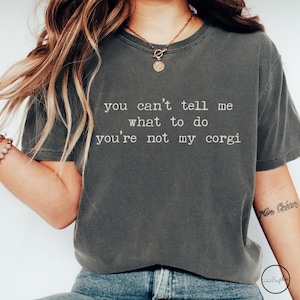 May include: Dark gray t-shirt with the text "you can't tell me what to do you're not my corgi" in white. The shirt has short sleeves and a crew neck. A gold necklace with a pendant is visible.