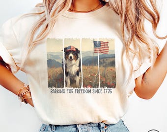 May include: Cream-colored t-shirt featuring a graphic of a dog wearing an American flag hat, with an American flag in the background. The text "BARKING FOR FREEDOM SINCE 1776" is printed below the graphic. The shirt is made of a soft material.