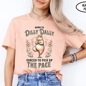 May include: A woman wearing a heather peach t-shirt with the text "Born to Dilly Dally, Forced to Pick Up The Pace" and a graphic of a capybara doing yoga.