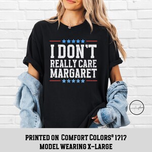 May include: Black t-shirt with white text that reads "I don't really care Margaret" with red and white stars above and below the text. The shirt is printed on Comfort Colors&reg; 1212 and the model is wearing a size X-Large.