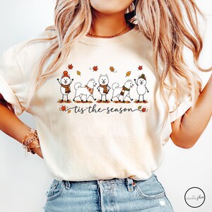 May include: A cream-colored t-shirt featuring a graphic of five white dogs wearing autumn-themed scarves and hats, surrounded by falling leaves. Below the dogs, the text reads 'tis the season' in a black cursive script font.