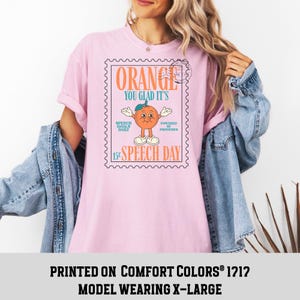 Puede incluir: Camiseta rosa claro con un dise&ntilde;o gr&aacute;fico de una naranja en un dise&ntilde;o de sello postal. El texto en la camiseta dice "ORANGE YOU GLAD IT'S SPEECH DAY". La camiseta es un modelo Comfort Colors 1212, talla X-Large.