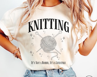 May include: A white t-shirt with black text that reads "Knitting It's Not A Hobby, It's A Lifestyle". The shirt features a black and white illustration of a ball of yarn with two knitting needles.