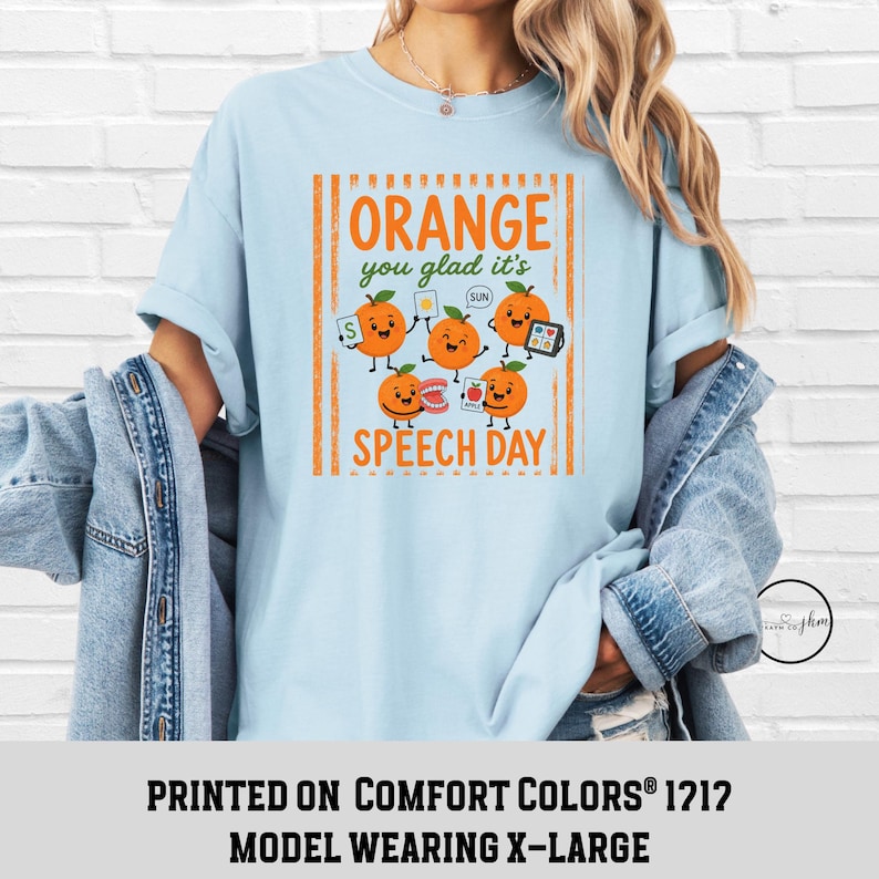 Pu&ograve; includere: Una t-shirt azzurro chiaro con una grafica di cinque arance cartoon con volti. Il testo recita "ORANGE you glad it's SPEECH DAY". Le arance tengono in mano strumenti di logopedia, tra cui flashcard, un modello di denti e una tavola di comunicazione.