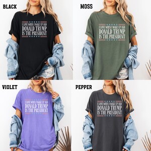Op de afbeelding: Vier T-shirts in verschillende kleuren met de tekst "Ik hou ervan om wakker te worden en Donald Trump is president" erop gedrukt. De tekst is wit met rode en blauwe sterren.