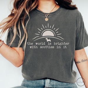 May include: Dark gray t-shirt with a white graphic of a sun, a Scottie dog, and the text "the world is brighter with scotties in it." The shirt has a crew neck and short sleeves.