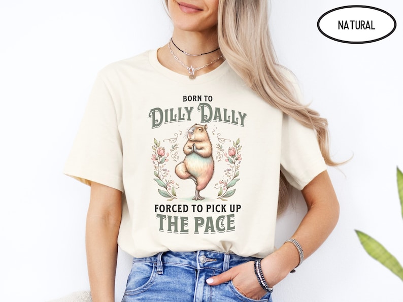 May include: A cream-coloured t-shirt with the text "Born to Dilly Dally, Forced to Pick Up The Pace" in a vintage style font. A cartoon capybara is doing yoga in the centre of the shirt.
