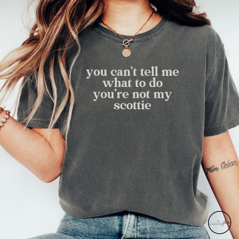 Puede incluir: Camiseta gris oscuro con el texto blanco "you can't tell me what to do you're not my scottie". La camiseta tiene mangas cortas y cuello redondo. Se ve un collar dorado con un colgante.