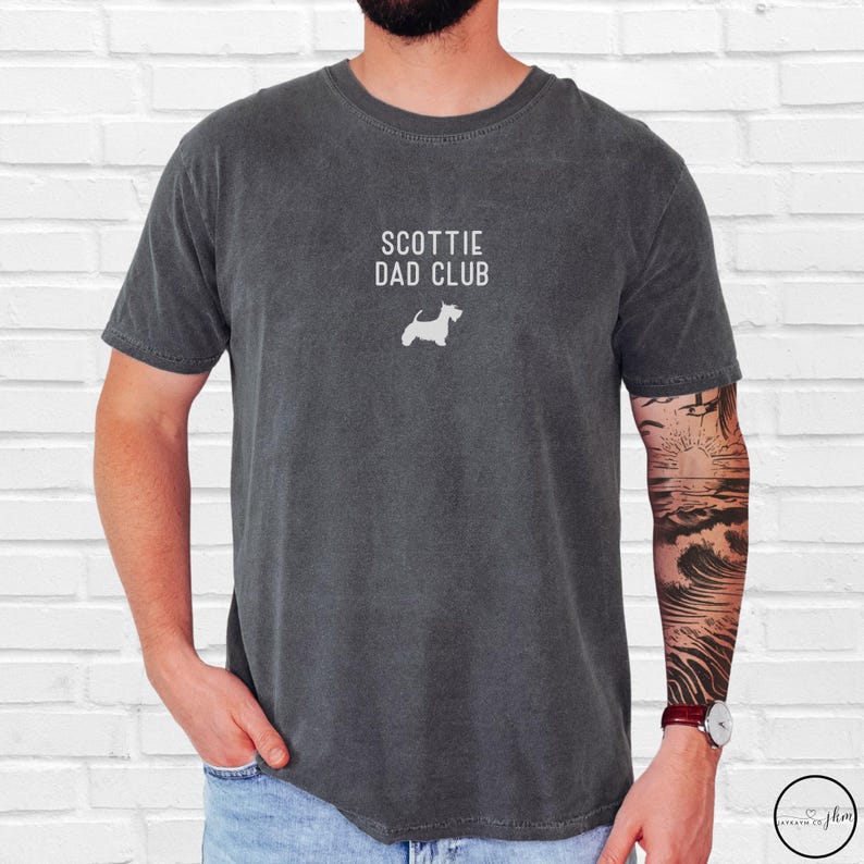 May include: Dark gray t-shirt with the text "SCOTTIE DAD CLUB" and a white silhouette of a Scottish Terrier. The shirt is a crew neck and has a relaxed fit.