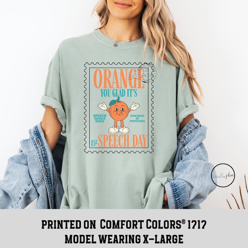 Puede incluir: Camiseta verde claro con un dise&ntilde;o retro de una naranja sonriente y el texto "ORANGE YOU GLAD IT'S SPEECH DAY". La camiseta tambi&eacute;n tiene el texto "SPEECH GOALS DAILY" y "POWERED BY PHONEMES".