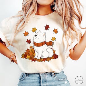 May include: A cream-colored t-shirt featuring a graphic of a white fluffy dog wearing an orange and brown plaid scarf, sitting in a pile of autumn leaves with additional maple leaves floating in the air around the dog.