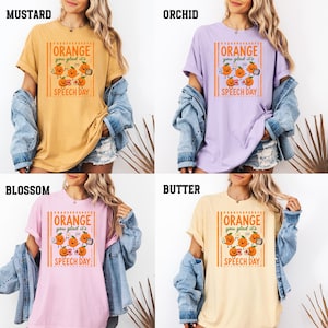 Pu&ograve; includere: Una collezione di quattro magliette a maniche corte nei colori senape, orchidea, fiore e burro. Ogni maglietta presenta una stampa grafica di arance cartoon con il testo "ORANGE you glad it's SPEECH DAY" all'interno di una cornice rettangolare.