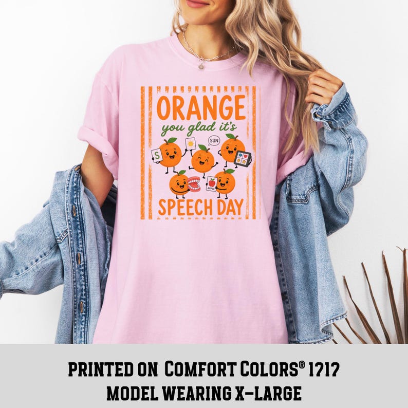 Pu&ograve; includere: Una t-shirt rosa chiaro con una grafica di cinque arance cartoon con braccia e gambe. Il testo recita "Orange you glad it's SPEECH DAY" in lettere arancioni. Le arance tengono in mano oggetti di logopedia come una carta con la S, un sole, denti e un tablet.