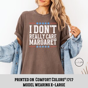 May include: A brown t-shirt with the text "I DON'T REALLY CARE MARGARET" printed in white. The text is surrounded by red and blue stars. The shirt is being modeled by a person wearing a denim jacket.