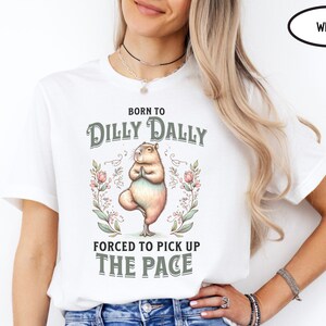 May include: White t-shirt featuring a graphic of a capybara doing yoga with the text "Born to Dilly Dally Forced to Pick Up The Pace" and floral accents.