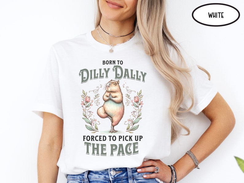 May include: White t-shirt featuring a graphic of a capybara doing yoga with the text "Born to Dilly Dally Forced to Pick Up The Pace" and floral accents.