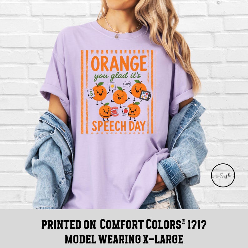 Pu&ograve; includere: Una t-shirt viola chiaro con una grafica di cinque arance cartoon con volti. Il testo recita "ORANGE you glad it's SPEECH DAY". Le arance tengono in mano vari oggetti, tra cui una carta con la lettera S, una carta sole, una dentiera e una carta mela.