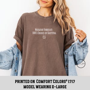May include: A brown t-shirt with the text "Weekend Forecast: 100% Chance of Knitting" and a small illustration of a ball of yarn and knitting needles. The model is wearing a size X-Large. The shirt is printed on Comfort Colors&reg; 1212.